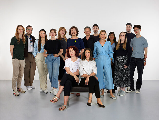
            
                a group of people in front of a white wall, four of them sitting on a bench in the foreground: the marketing and communication team at mumok
            
        