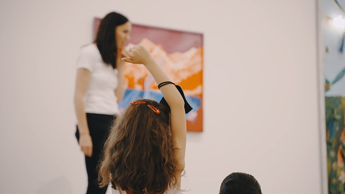 ein Kind mit langen Haaren zeigt auf, es hat eine rote Masche im Haar. Unscharf im Hintergrund eine junge Frau mit dunklen Haaren und weißem Shirt vor einem bunten Kunstwerk