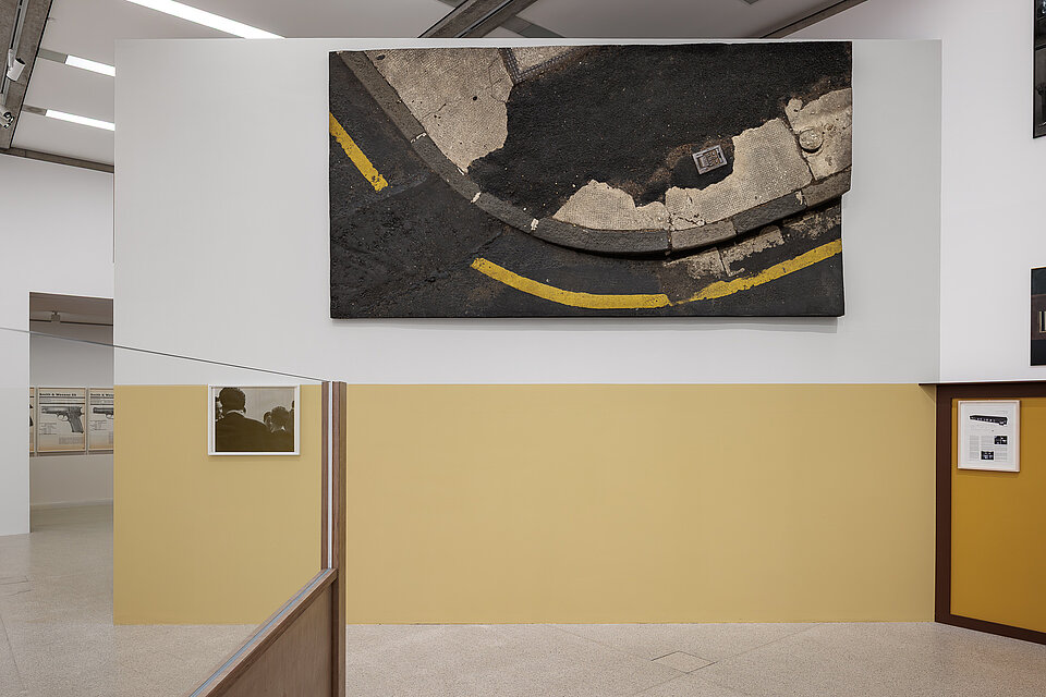 
            
                Ausstellungsansicht mit einer großformatigen Bildarbeit, die eine Straßenoberfläche mit gelben Fahrbahnmarkierungen zeigt; darunter und seitlich befinden sich gerahmte Fotografien und Dokumente an einer zweifarbigen Wand.
            
        