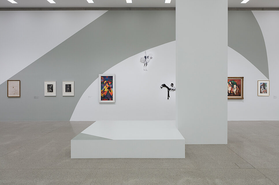 A large white-grey pedestal in the foreground. Behind it hang six pictures and two sculptures on the walls.