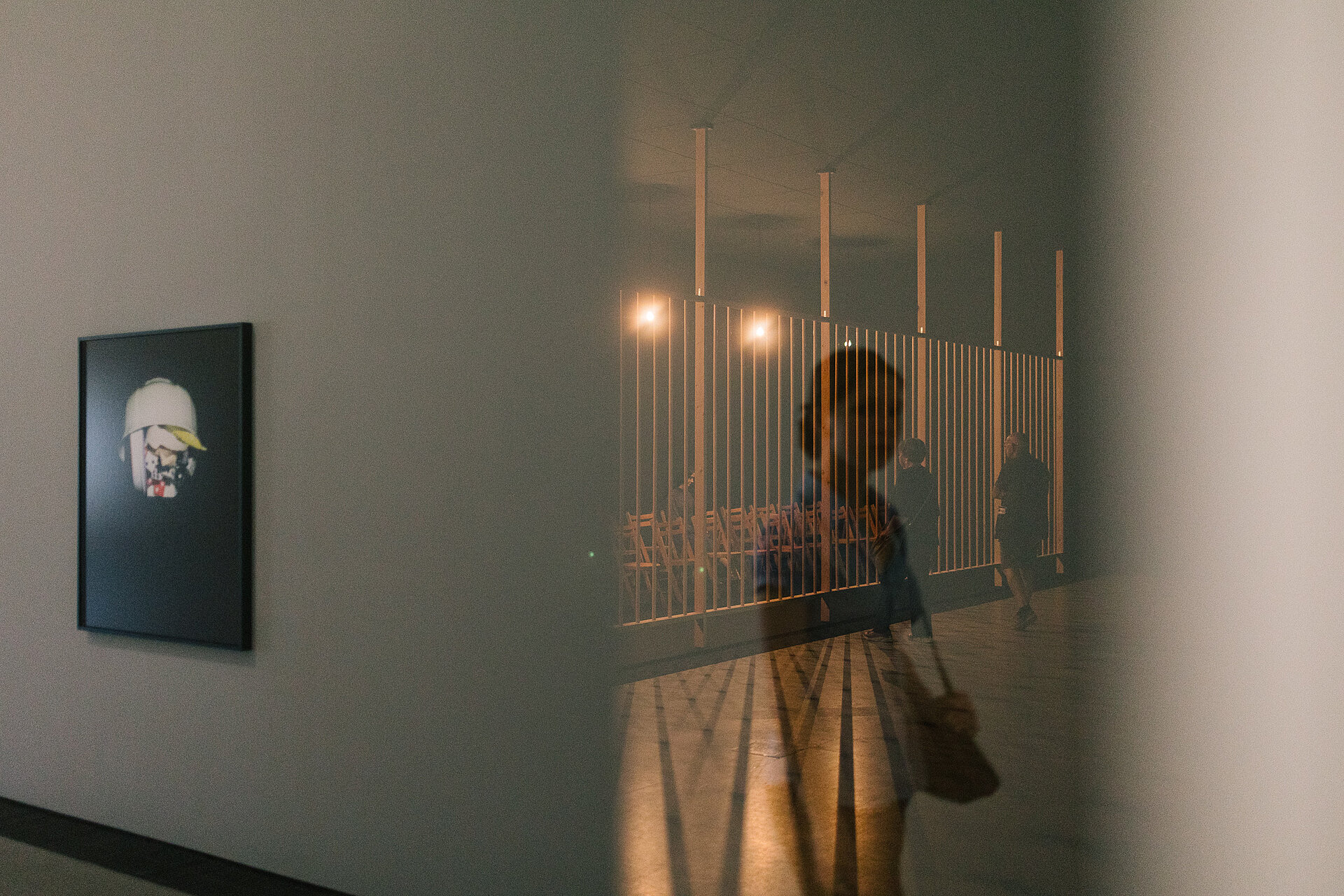 
            
                Blick in einen Ausstellungsraum mit gerahmter Fotografie an der Wand; durch eine spiegelnde oder transparente Fläche sind Besucher:innen sowie eine Installation aus vertikalen Stäben und Sitzreihen sichtbar. / View into an exhibition space with a framed photograph on the wall; reflected or seen through a transparent surface are visitors and an installation consisting of vertical rods and rows of seating.
            
        
