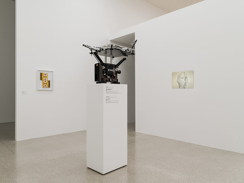 A base in the exhibition space with a large projector projecting an image of a head onto the wall on the right and framed video tapes in the background on the left