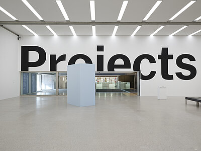 [ID: A sculpture of oversized black letters painted onto white museum walls. From this view, the letters spell “Proiects,” with the “j” of “Projects” appearing as an “i:” some letters are cut off by the gallery’s doorways and corners. On the image’s right sits a small sculpture on a white plinth. In the image’s center, another rectangular sculpture, much larger and made of pastel blue polyurethane foam, partially obscures the gallery’s glass entrance and view into the museum’s lobby.]