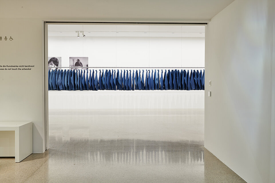 Blick in einen Ausstellungsraum, in dem an einer langen Stange viele blaue Hemden hängen