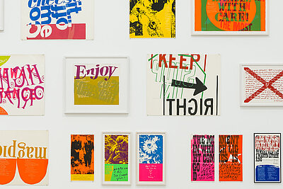 weiße Wand mit knapp 15 kleinen Grafiken und Gemälden, manche davon zeigen Schriftzüge, eines etwa den Ausstellungstitel Enjoy