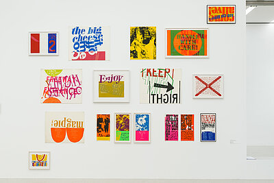 white wall with almost 15 small graphics and paintings, some of which show lettering, such as the exhibition title Enjoy