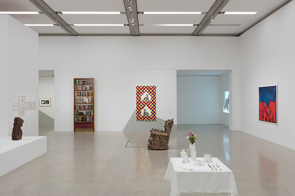  On the right, a picture with two red animals on a blue background. In the centre, two sculptures in the form of a laid table and a reading chair. Behind them are two further works of art. On the left, a sculpture on a base. 