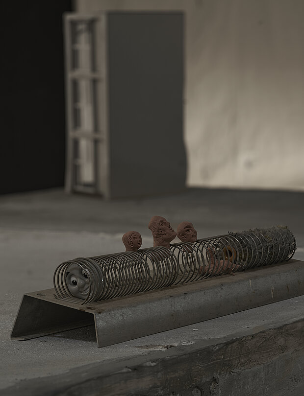 
            
                Skulpturale Installation auf einem Betonsockel mit mehreren Metallspiralen, aus denen kleine keramische Kopf- und Figurenfragmente hervorragen; am linken Ende einer Spirale ist ein stilisiertes Gesicht sichtbar. Im Hintergrund befinden sich ein Ausstellungsraum mit Betonboden und eine unscharf sichtbare, freistehende Wandstruktur. / Sculptural installation on a concrete plinth consisting of several metal coils from which small ceramic heads and figure fragments emerge; a stylized face is visible at the left end of one coil. In the background are an exhibition space with a concrete floor and a blurred freestanding wall structure.
            
        