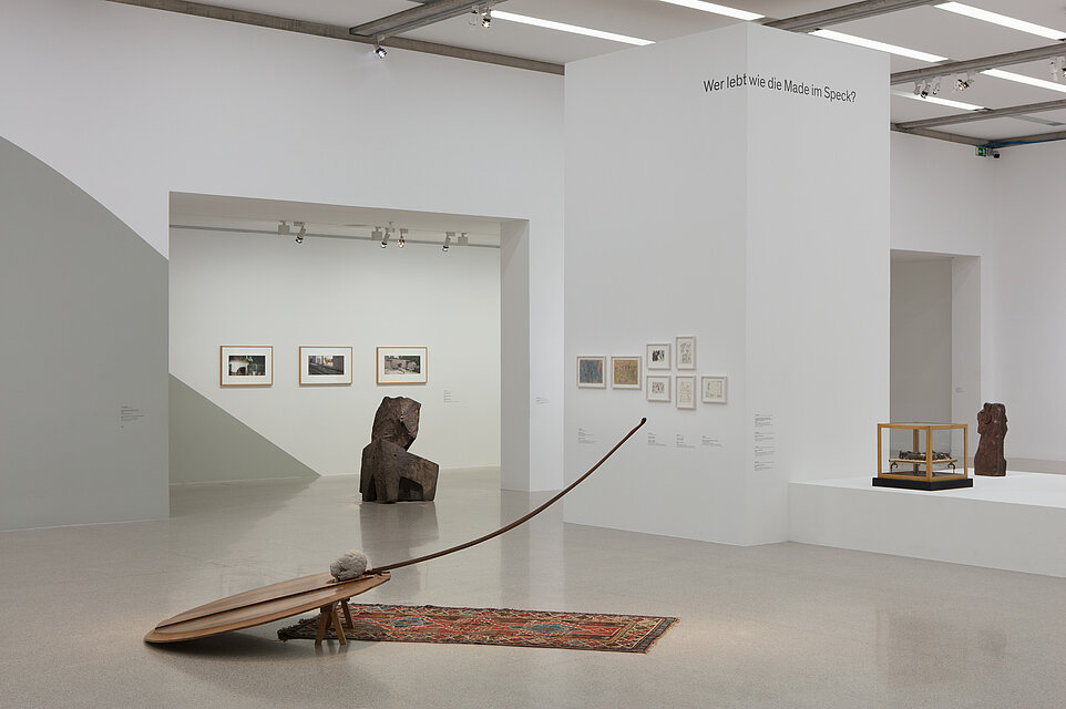 In the foreground, a sculpture made of wood and a colourful carpet on the floor. Behind it, an abstract metal sculpture. In the background, three framed photos and seven other small-format pictures. 