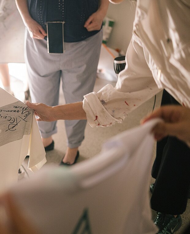 
            
                Hände halten bedruckte Stoffstücke oder Kleidungsstücke, während im Hintergrund weitere Personen stehen und Materialien betrachten. / Interior scene with several people: hands hold printed fabric pieces or garments while additional people stand in the background examining materials.
            
        