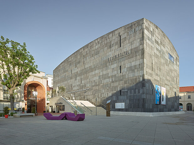 
            
                Außenansicht eines massiven Museumsgebäudes mit grauer Steinfassade und dem Schriftzug „mumok“, davor ein offener Platz mit Treppen, Sitzmöbeln und angrenzenden historischen Gebäuden. / Exterior view of a large museum building with a grey stone facade labeled “mumok,” featuring an open square with stairs, seating elements, and adjacent historic buildings.
            
        