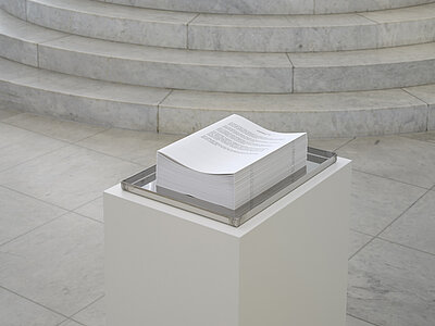 Park McArthur, Contact M, 2025, [ID: Ein hoher Stapel von Broschüren im Briefformat wird in einem flachen Tablett aus Edelstahl angeboten. Das Tablett ruht auf einem weißen Sockel in einem Galerieraum mit einer geschwungenen Marmortreppe im Hintergrund.] 