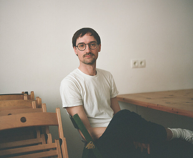 
            
                Philipp Fleishcmann sitzend an einem Tisch vor einer weißen Wand, er trägt ein weißes Tshirt und eine Brille
            
        