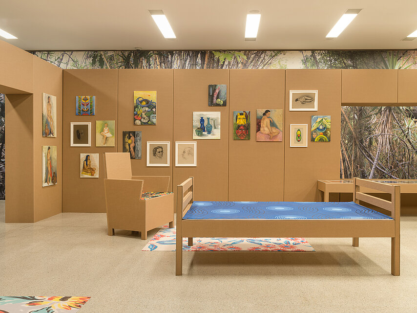 Reconstruction of a cardboard living room, in the middle a cardboard bed with a blue cover, in the background colourful works of art on the wall