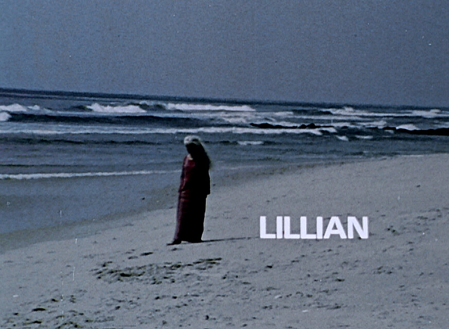 Filmbild einer einzelnen Person an einem Sandstrand vor dem Meer; im rechten unteren Bereich steht der eingeblendete Schriftzug „LILLIAN“. / Film still showing a single person standing on a sandy beach facing the sea; the text “LILLIAN” appears in the lower right area of the image.