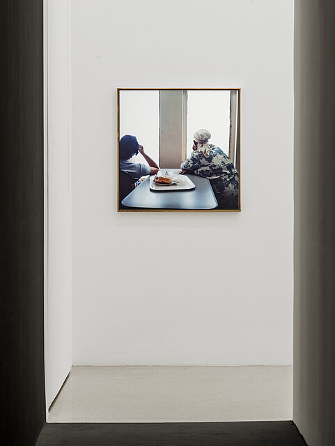 Fotografie von zwei Menschen, die aus einem Fenster blickend am Tisch sitzen, die Fotografie hängt an einer weißen Wand