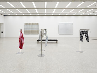 [ID: A wide gallery view of multiple artworks, including two large sculptures made of unadorned American and Austrian highway signs. These sculptures resemble one another in their dimension, composition, and softly reflective aluminum surfaces. To the left, a rubber loading dock bumper is bolted to the gallery wall. In the foreground sit two dark leather museum benches and four sculptures made of tall metal stands from which torn pajama pants of various patterns, colors, and fabrics hang.]
