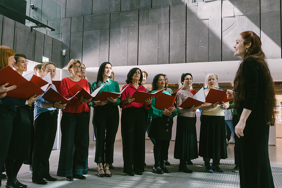 Gruppe von Personen mit roten Chormappen in einem modernen Innenraum mit hohen Betonwänden; im Vordergrund steht eine dirigierende Person vor dem Halbkreis des Chors. / Group of people holding red choir folders in a modern interior with high concrete walls; in the foreground, a conductor stands facing the semicircle of singers.