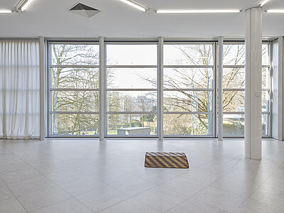 Park McArthur, Untitled, 2014, [ID: Eine kleine Rampe mit einem gelb-rot gestreiften Muster, die direkt auf dem Boden der Galerie liegt. Vor großen Fenstern platziert, steht die Rampe im Kontrast zur umgebenden Architektur. Natürliches Licht erfüllt den Raum, im Hintergrund sind Bäume und eine Stadtlandschaft durch das Glas zu sehen.]