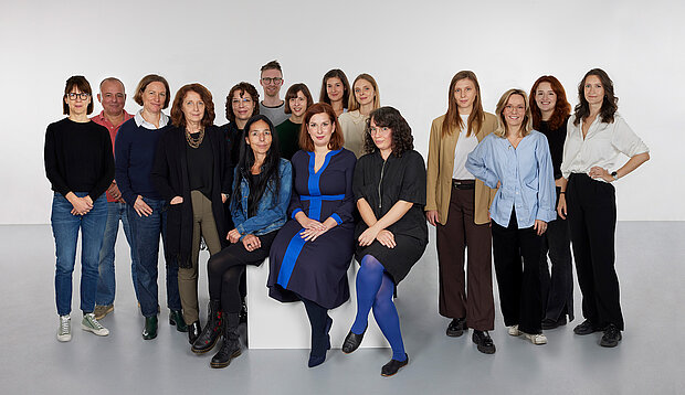 
            
                eine Gruppe von Personen vor einer weißen Wand, drei davon sitzen im Vordergrund auf einem Sockel, das Team Sammlung im mumok
            
        