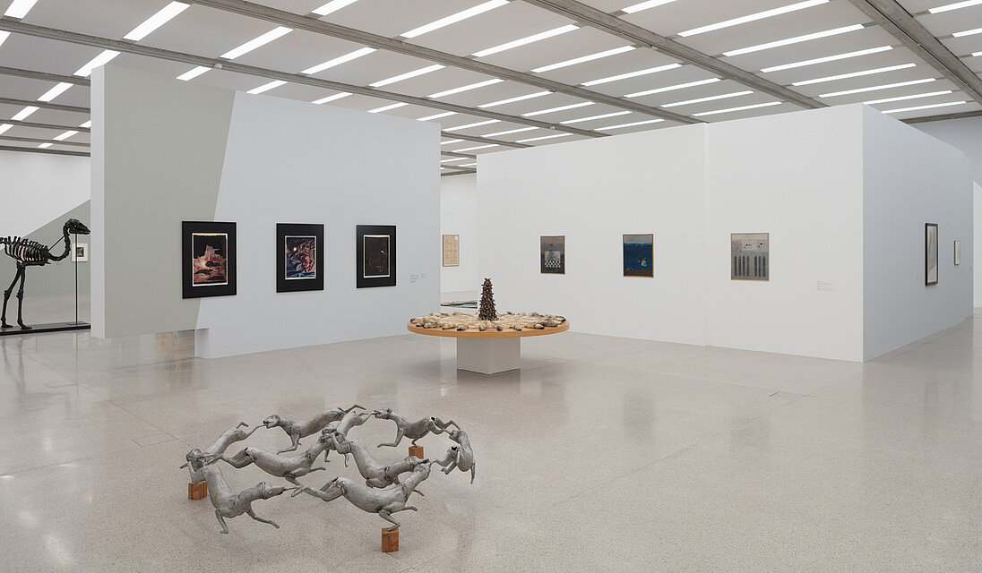 Im Ausstellungsraum zentral zwei Skulpturen: vorne eine Plastik aus Metallhunden, dahinter ein massiver runder Holztisch mit Fellteilen von Tieren darauf. Im Hintergrund diverse Bilder an den Wänden.