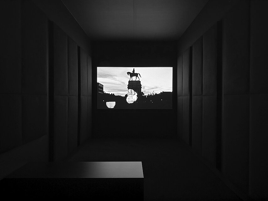  View into a dark room with dark curtains at the sides, in the centre is a screen with a black and white film showing a statue of an equestrian