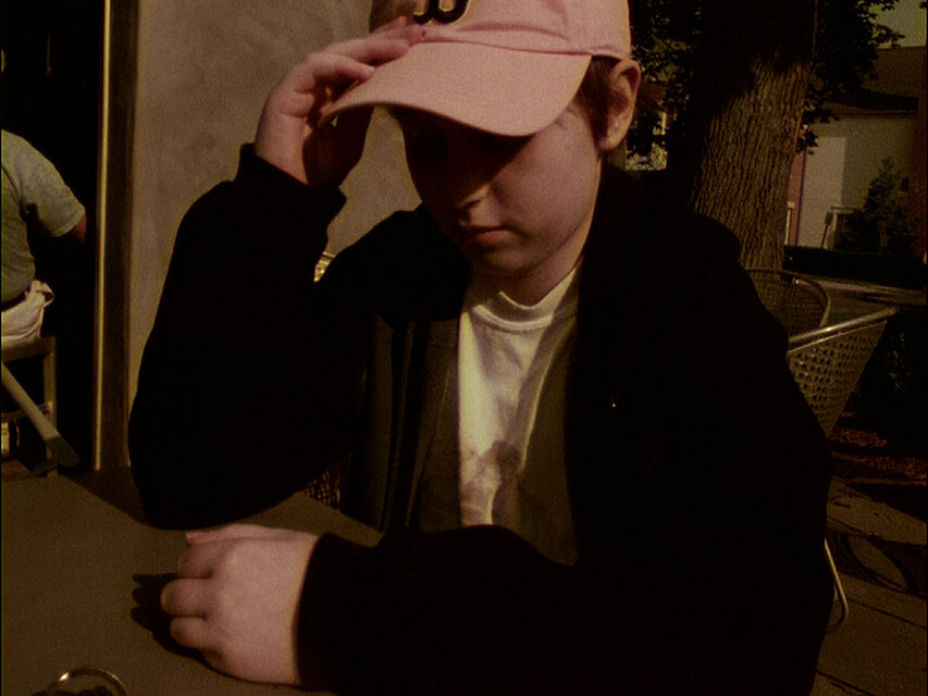 Person mit Baseballkappe sitzt an einem Tisch im Freien im warmen Abendlicht. / Person wearing a baseball cap sitting at an outdoor table in warm evening light.