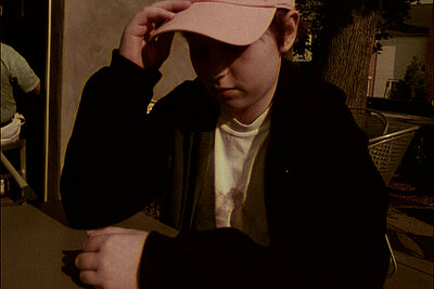 Person mit Baseballkappe sitzt an einem Tisch im Freien im warmen Abendlicht. / Person wearing a baseball cap sitting at an outdoor table in warm evening light.