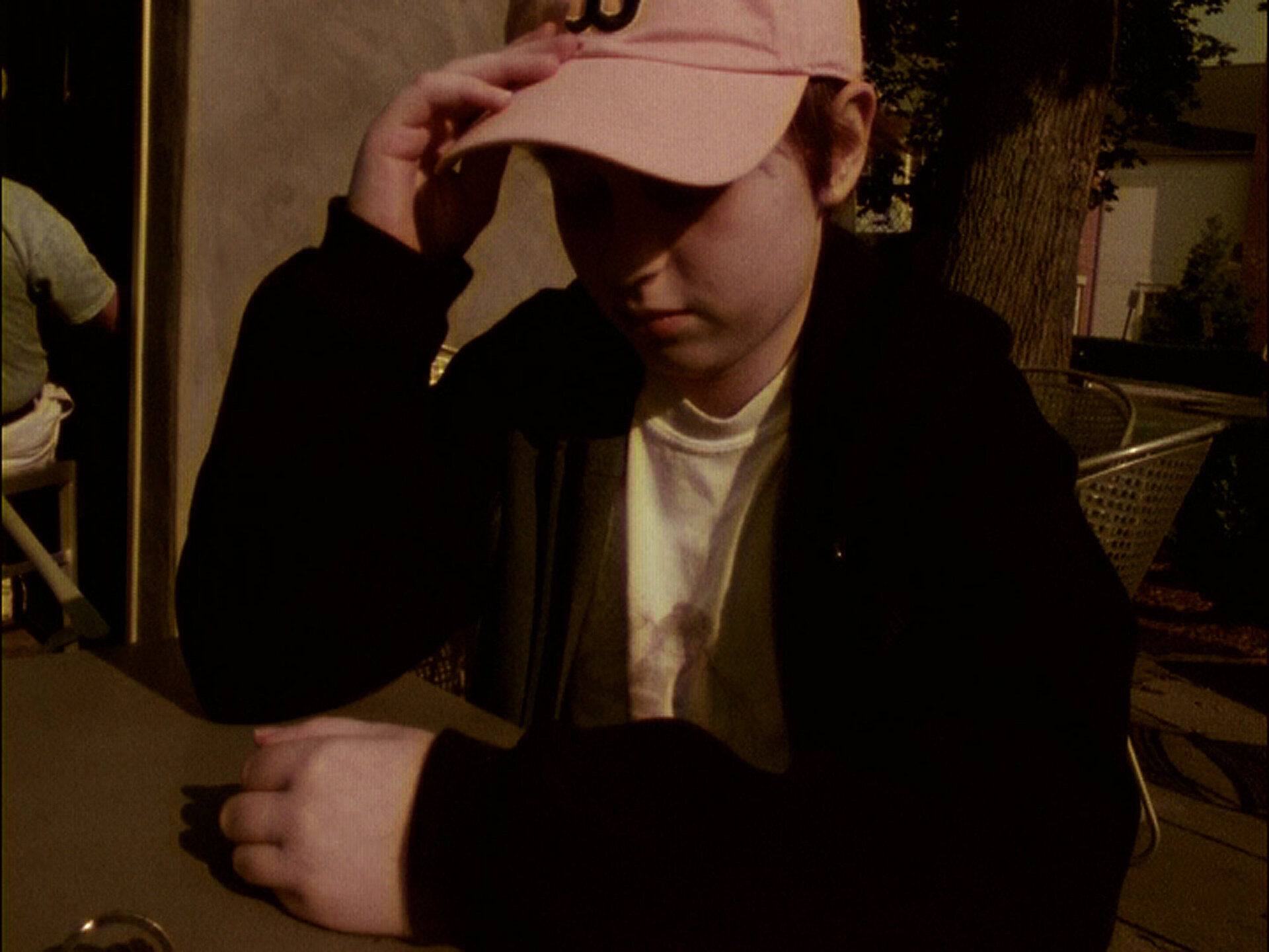Person mit Baseballkappe sitzt an einem Tisch im Freien im warmen Abendlicht. / Person wearing a baseball cap sitting at an outdoor table in warm evening light.