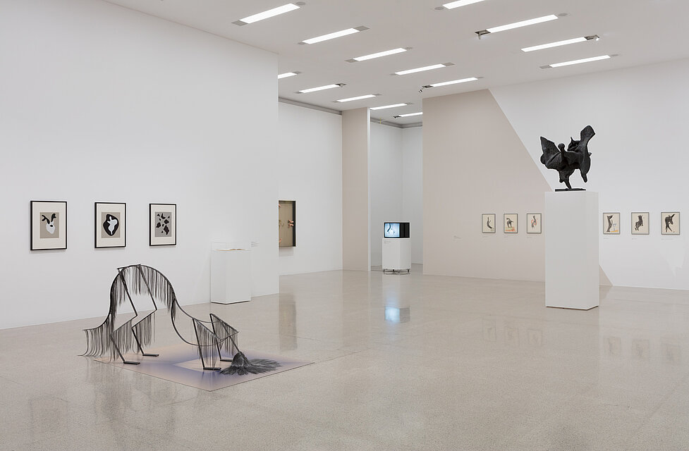 In the foreground, two sculptures in abstract animal shapes. A tube television is placed on a pedestal in the background. Small-format paintings hang on the walls to the right and left of it.