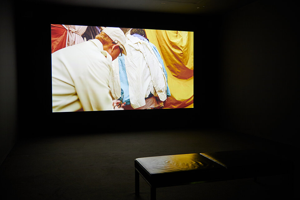 dark room, central screen showing a film, a colourful picture with white and yellow textiles, a man cuts something off the textiles