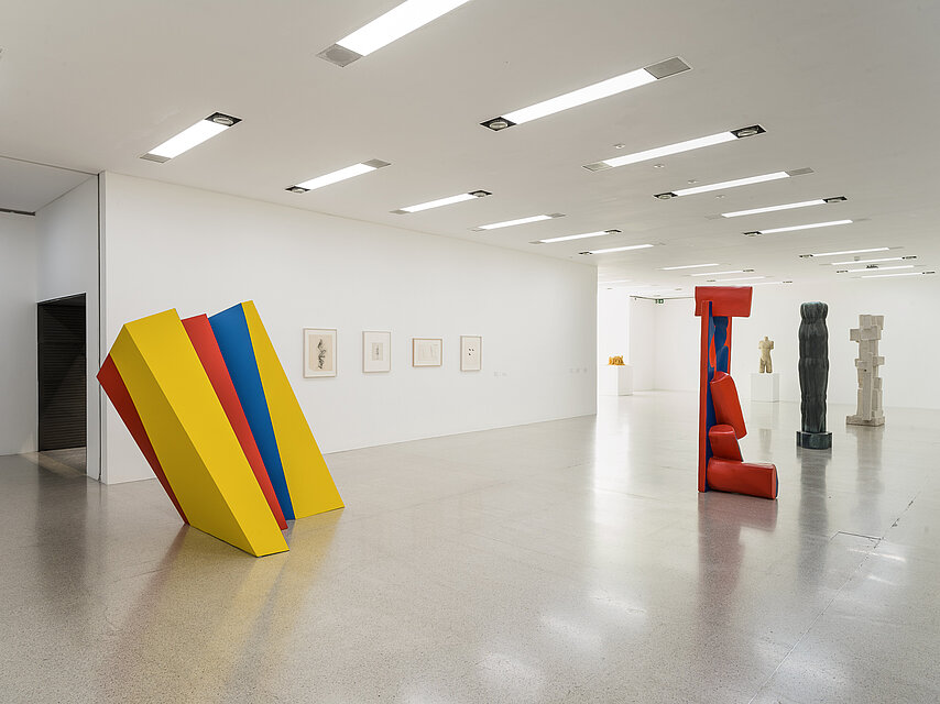  View of a bright exhibition space, on the left a geometric sculpture in yellow, red and blue, on the right a red sculpture in the background