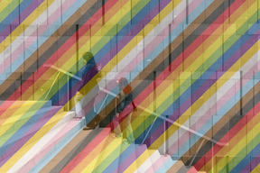 drei junge Frauen gehen die Treppen vor der mumok Fassade hinauf und berühren die Wand, auf dem Bild ist ein leicht transparenter Regenbogen zu sehen