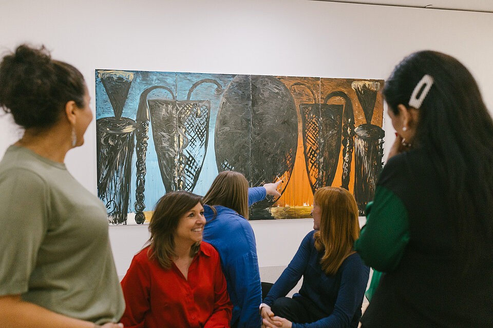 Mehrere Personen befinden sich vor einem großformatigen Gemälde mit schwarzen Formen auf blauem und orangefarbenem Hintergrund; eine Person zeigt auf das Kunstwerk, während andere davor sitzen oder stehen. / Several people are gathered in front of a large painting with black forms on blue and orange backgrounds; one person points toward the artwork while others are seated or standing nearby.