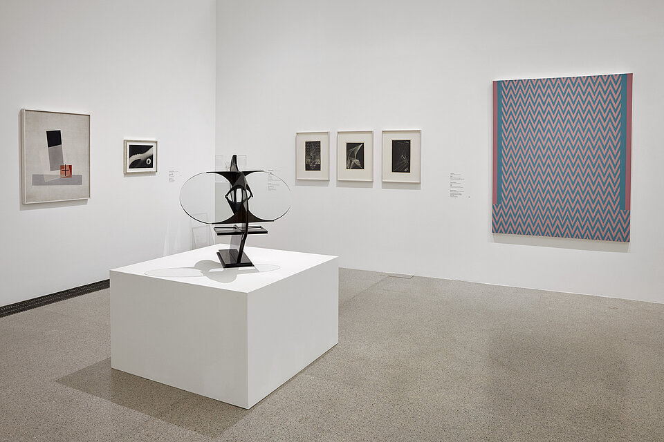 a black, filigree sculpture stands on a white base in the centre of the picture, with pictures of different sizes in dark colours on white walls in the background