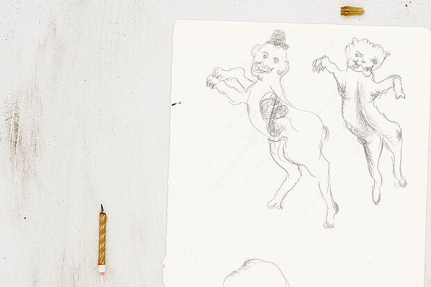 
            
                Papier mit Zeichnungen zweier aufrecht stehender, tierähnlicher Figuren, links daneben eine kurze Kerzenstummel auf der Oberfläche. / Sheet of paper showing drawings of two upright, animal-like figures, with a short candle stub placed to the left.
            
        