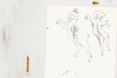 Papier mit Zeichnungen zweier aufrecht stehender, tierähnlicher Figuren, links daneben eine kurze Kerzenstummel auf der Oberfläche. / Sheet of paper showing drawings of two upright, animal-like figures, with a short candle stub placed to the left.