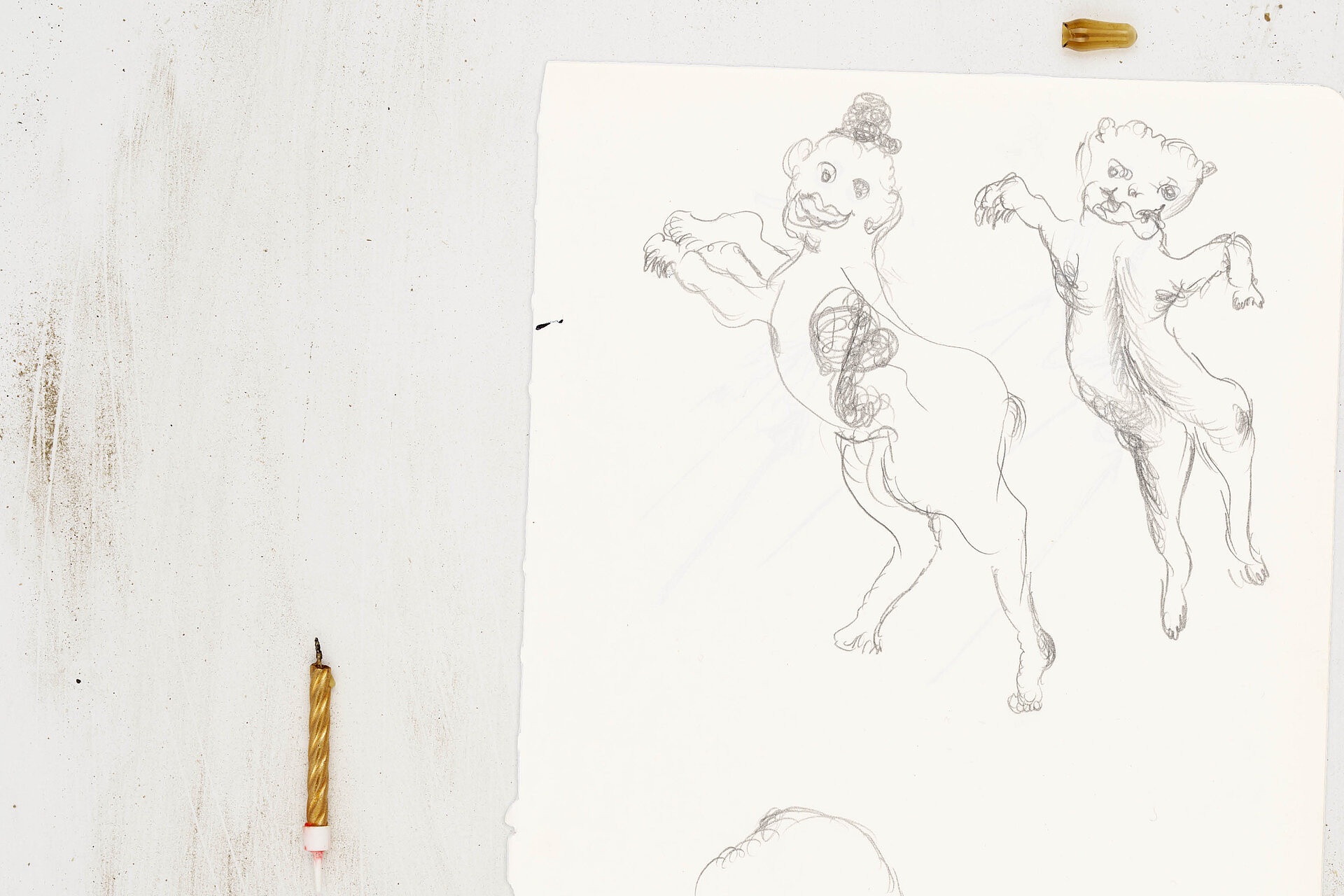 Papier mit Zeichnungen zweier aufrecht stehender, tierähnlicher Figuren, links daneben eine kurze Kerzenstummel auf der Oberfläche. / Sheet of paper showing drawings of two upright, animal-like figures, with a short candle stub placed to the left.