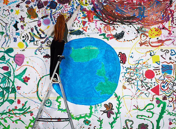 
            
                Eine Frau steht auf einer Leiter vor einem großen Bunten Bild, das einen Erdball und bunte Dekorationen zeigt
            
        