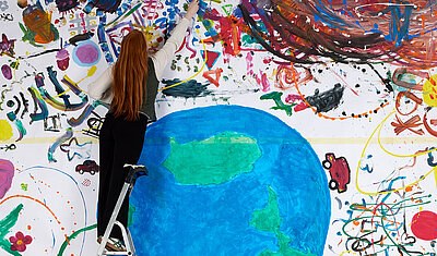 Eine Frau steht auf einer Leiter vor einem großen Bunten Bild, das einen Erdball und bunte Dekorationen zeigt