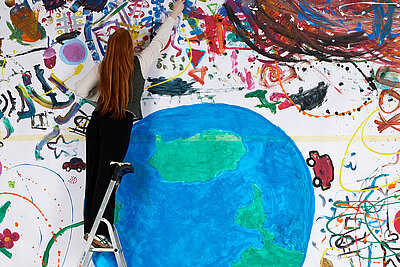 Eine Frau steht auf einer Leiter vor einem großen Bunten Bild, das einen Erdball und bunte Dekorationen zeigt