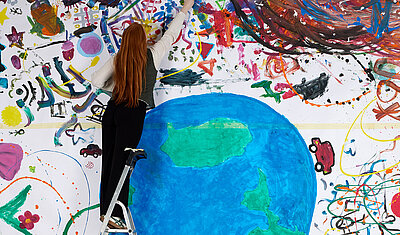 Eine Frau steht auf einer Leiter vor einem großen Bunten Bild, das einen Erdball und bunte Dekorationen zeigt