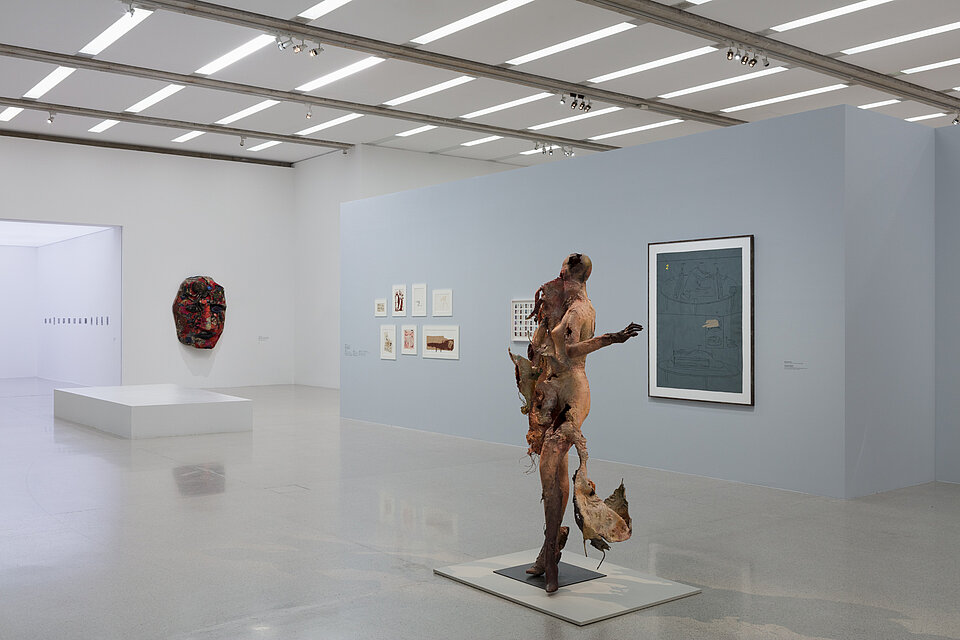 An abstract figurative sculpture in the foreground. Behind it, works of art on the wall. In the background, an oversized red face on the wall. 