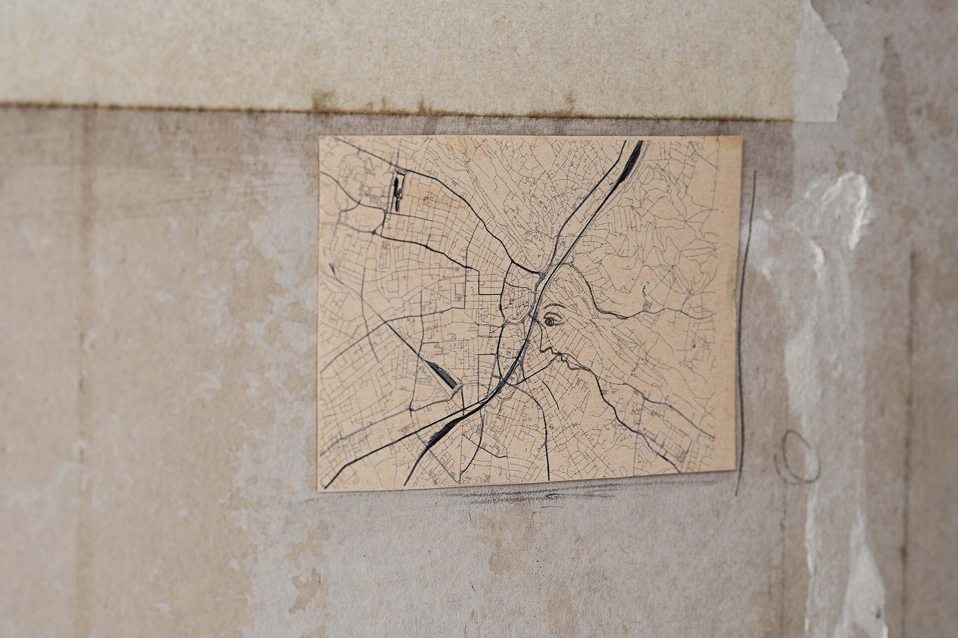 Kleine rechteckige Zeichnung auf gealtertem Papier, die ein dichtes Netz aus kartografischen Linien zeigt, in das das Profil eines menschlichen Gesichts eingezeichnet ist. / Small rectangular drawing on aged paper showing a dense network of cartographic lines with the profile of a human face integrated into the map structure.