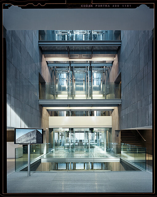 Symmetrische Innenansicht eines modernen Gebäudes mit mehreren gläsernen Aufzügen, metallischen Strukturen und Etagen, eingerahmt von Betonwänden. / Symmetrical interior view of a modern building with multiple glass elevators, metal structures, and floors framed by concrete walls.
