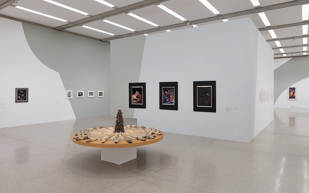 In the centre of the room is a solid, round wooden table. Pieces of animal fur lie on it. Various paintings and photographs hang on the walls in the background.