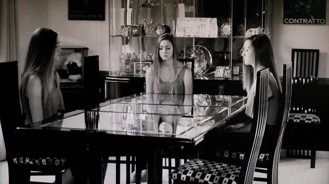 A black-and-white photograph of a reflective table with three identical seated figures shown frontally and through reflections. The room includes display cabinets, chairs, and decorative objects in the background