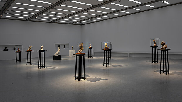 
            
                Großer Ausstellungsraum mit mehreren Skulpturen und Büsten auf hohen dunklen Sockeln, gleichmäßig im Raum verteilt und von Oberlichtern beleuchtet. / Large exhibition gallery with multiple sculptures and busts displayed on tall dark pedestals, evenly distributed throughout the space and illuminated by overhead skylights.
            
        