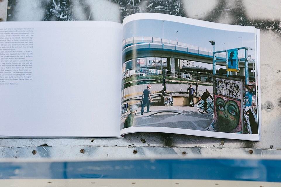 Aufgeschlagenes Fotobuch auf einer rauen Metall- oder Steinoberfläche, die rechte Seite zeigt eine urbane Szene unter einer Hochstraße mit Personen, Fahrrädern und Graffiti, die linke Seite enthält einen Textblock. / An open photo book placed on a rough metal or stone surface, with the right page showing an urban scene beneath an overpass with people, bicycles, and graffiti, and the left page containing a block of text.