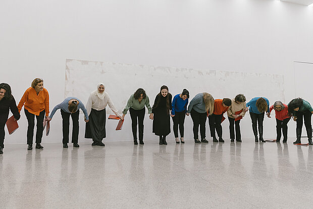 
            
                Frauen in bunten Pullovern stehen in einer Reihe, halten sich an den Händen und verbeugen sich
            
        